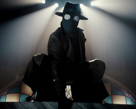 Nicolas Cage wearing black clothes and trilby and a face mask with spider-like eyes
