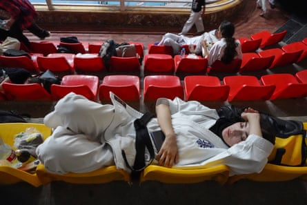 A woman in a gi with a black belt lies on plastic seats in a sports stadium.