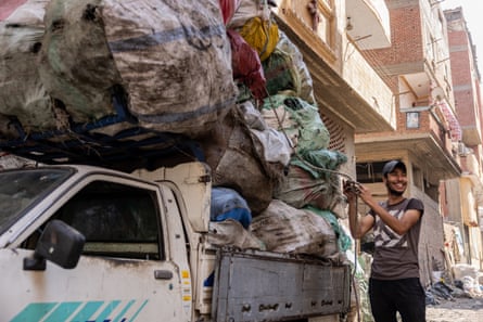 A smiling man uses rope to secure a towering pile of bagged rubbish on to the back of a pickup truck