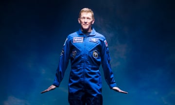 Tim Peake in a blue astronaut suit, behind him a dark blue night sky. His arms are by his side, hands flexed, as if he is taking off without a space craft.