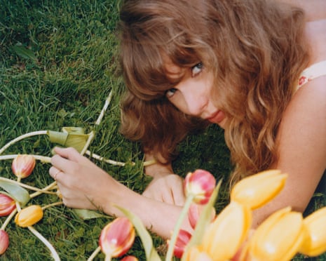 Taylor Swift with wavy hair lies on grass surrounded by yellow and pink tulips, looking up