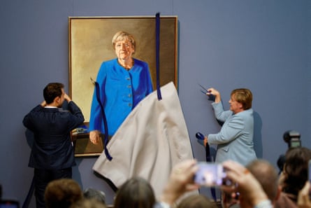 The portrait is unveiled at the museum: Merkel stands by holding a pair of scissors, having just cut a blue ribbon that held up a canvas covering, which is falling down. The artist stands to the side and people are taking photographs.