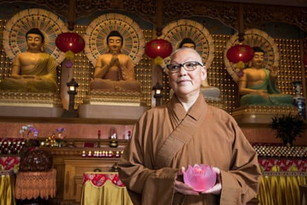 A Buddhist Venerable, dressed in robes and holding a moulded pink lotus, stands in front of a Buddhist shrine.