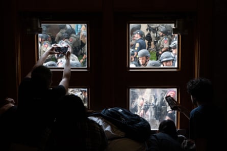 people gather inside a building, up against a window. through the window you can see many police in riot gear right nearby. one person in the building is holding up a phone to take a picture