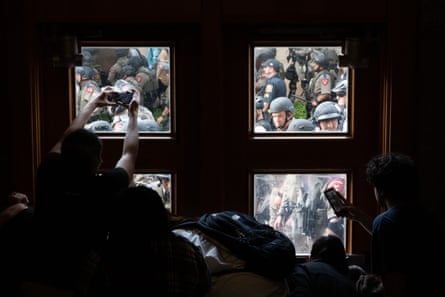 people gather inside a building, up against a window. through the window you can see many police in riot gear right nearby. one person in the building is holding up a phone to take a picture