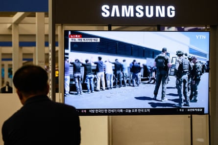 A man walks past a TV screen showing news clips of American men in combat gear with a line of Koreans facing a bus with their hands on it