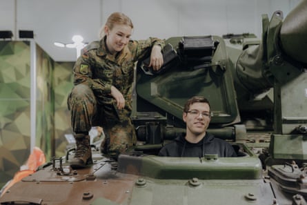A woman in military uniform squatting beside someone looking out from interior of tank