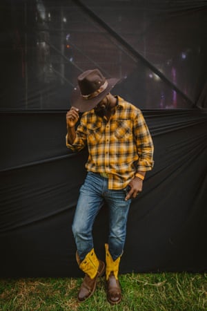 A man in yellow-checked western shirt, blue jeans and yellow and brown cowboy boots tips his hat