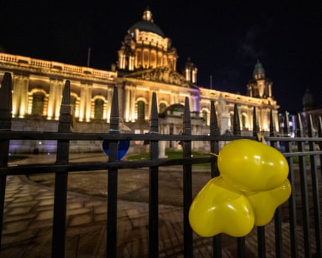 Yellow heart shaped balloons with the name shown of Jim Carson who passed away during the pandemic, are tied to the gates of Belfast City Hall which is illuminated in yellow to mark the National Day of Reflection.