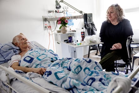 A man, with an oxygen tube in his nose, smiles as he lies under a duvet and looks at a woman with shoulder-length hair sitting next to his hospital bed, her hands in her lap and also smiling