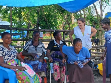 A woman in an MSF tabard stands talking to a group of seated women under a blue tarpaulin shelter.