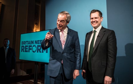 Farage and Jenrick stand on stage together. Jenrick is smiling while Farage is laughing and gesturing with one hand towards the audience. Farage wears a dark blue jacket, white shirt, and a pale pink tie with large dark spots on it.