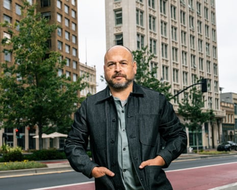 A bald man with a dark beard wears a dark-blue workwear-style jacket over a shirt, his hands in the pockets