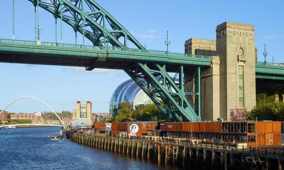 A row of red-brown shipping containers at the foot of a blue iron bridge on the banks of the river Tyne