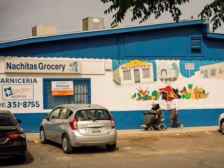 A man in an electric wheelchair and another man behind him walk in front of a blue and white small grocery store with signs that read “Nachitas Grocery” and with a mural depicting the original owners painted on the side