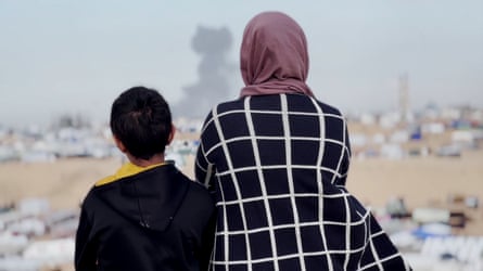 a boy in a hoodie and a woman in a coat and headscarf looking at a smoke coming from a destroyed-looking landscape