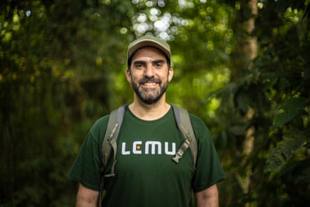 A man wearing a cap and a green T-shirt with the word Lemu stands in a forest.