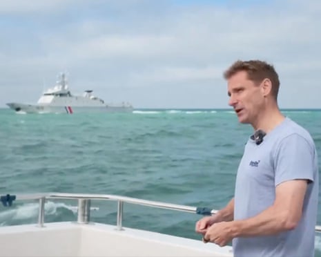 Chris Philp stands on a boat deck, with a warship visible on the water behind him