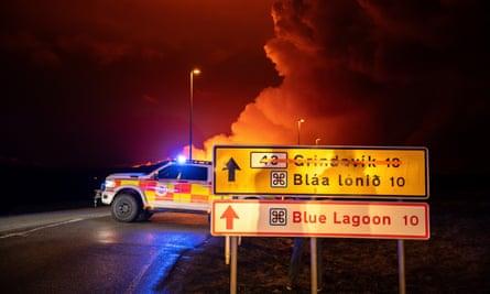 An emergency vehicle stationed on a road at night. An orange glow fills the sky and white smoke billows into the air. Two road signs are in the foreground, with Grindavik crossed out at the top