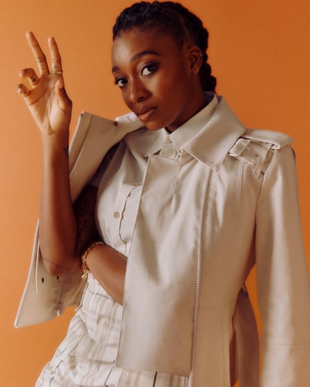Musician Little Simz, one hand up, in cream clothes against rust background