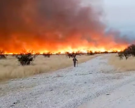 A person walking trhough scrubland with flames and clouds of smoke on the horizon