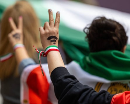 Two women draped in Iranian flags make the peace sign with the wifi symbol painted on the back of their hands.