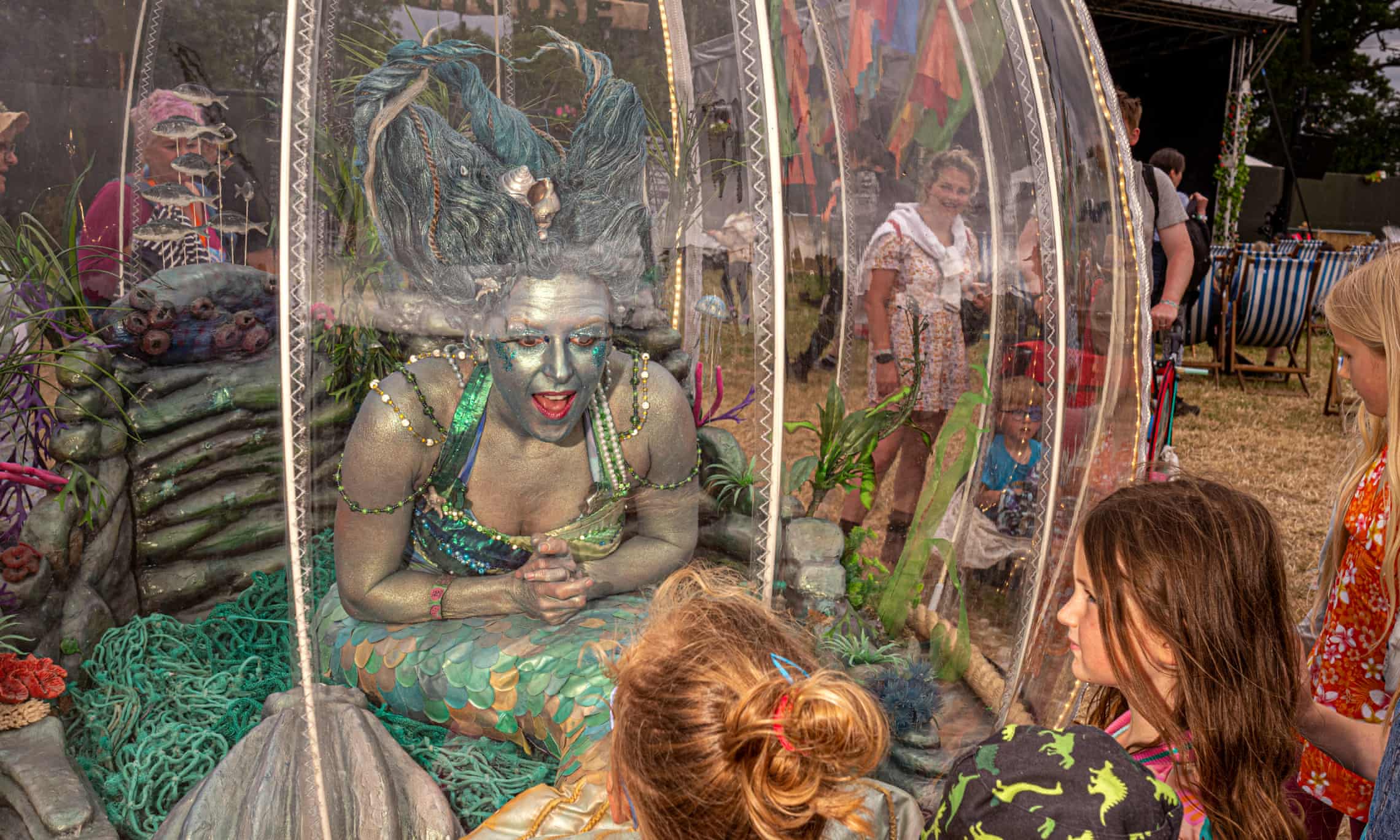 A woman dressed as a mermaid sits inside a large 'goldfish bowl' and talks to the children outside, watching her