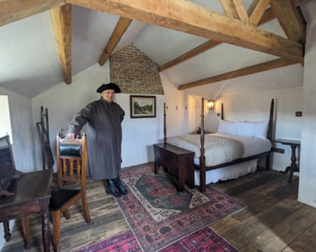 A man in period costume standing in a Georgian-style bedroom with a beamed ceiling, a simple four-poster bed and a writing desk