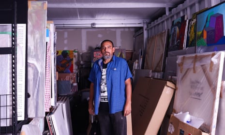 man wearing black shirt and open blue short-sleeve button-down surrounded by paintings