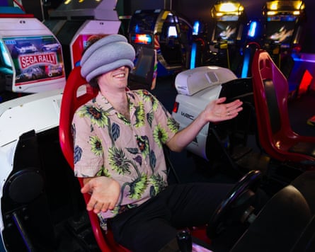 Pete Wise sits in the chair of an arcade car racing, wearing the Ostrichpillow over his eyes.