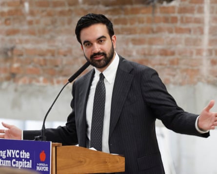 Zohran Mamdani gestures with arms spread wide from a lectern, with a bare-brick wall behind him.