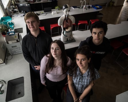 Four school students stand together in a science classroom, viewed from above, near an anatomical model