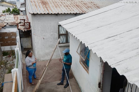 A woman and a man stand in a small courtyard next to a house with a white-painted roof