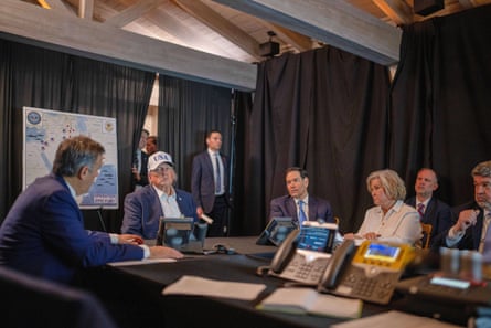 Donald Trump wearing a USA baseball cap sitting at a table with colleagues, with a map behind him