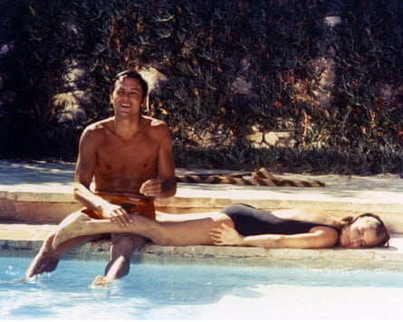A man whose lower legs dangle into a swimming pool puts his hand on the calf of a woman lying along the pool’s edge