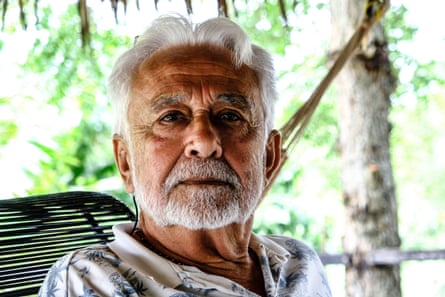 An older Hispanic man with white hair and a beard on a veranda looks at the camera