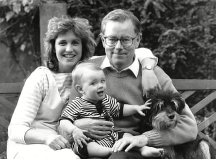 The parents are sitting on a wooden garden bench with Whitehall, on his father’s knee and stroking a dog that his father has his arm around.
