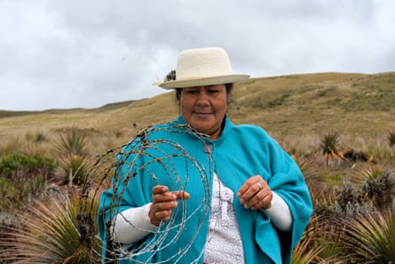 A woman in a blue shawl and white hat stands in a grassy plain holding a coil of barbed wire