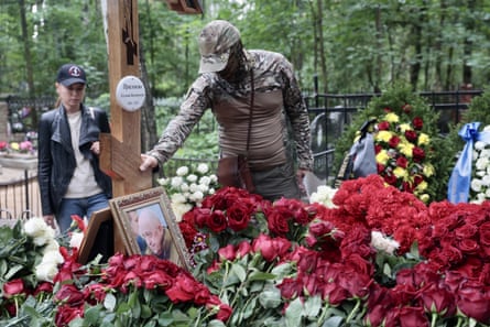 A man in military gear holds a wooden cross that has been placed on the grave of Prigozhin, which is laden with red flowers