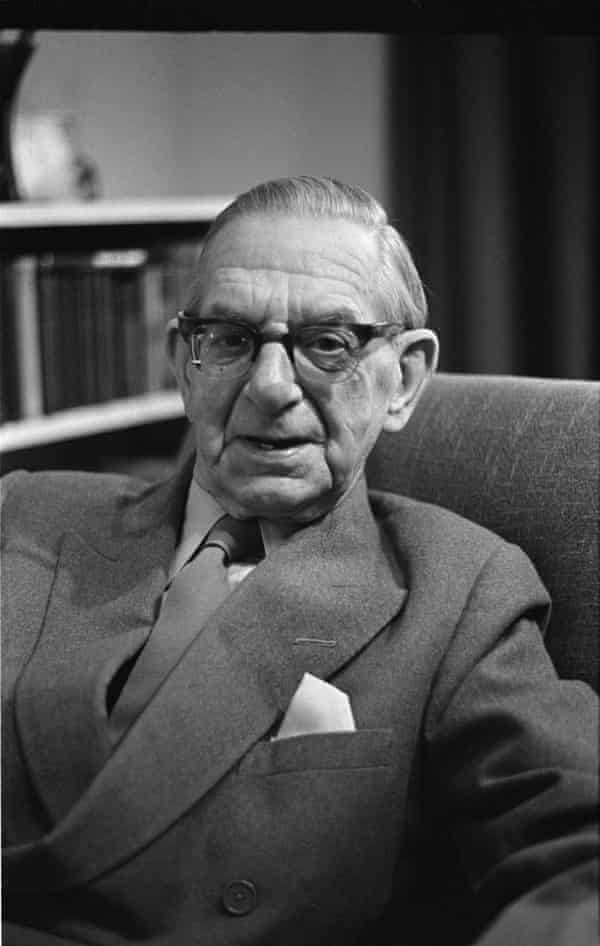 Neville Cardus in a suit and tie, reclining in an armchair with an out of focus bookcase behind him