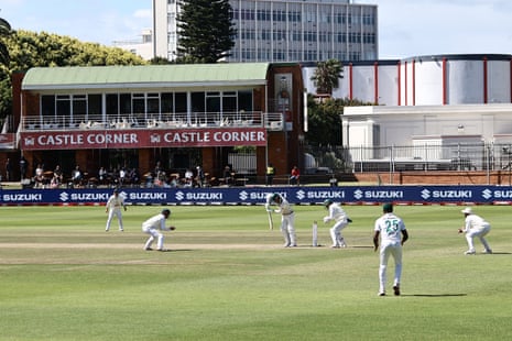 Cricketers in white uniforms play a Test match, with a batsman at the wicket near the pavilion