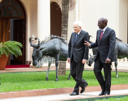 Two men in suits walk along a path past bird statues