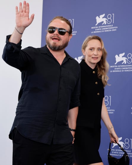 Brady Corbet, wearing black sunglasses, waves at a photo call as he holds the hand of Mona Fastvold