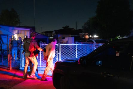 An officer escorts an arrested man through a gate at night, lit by red and blue lights.