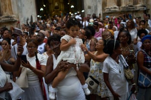 A child in a white dress is carried by a woman with a crowd of people in the street behind them