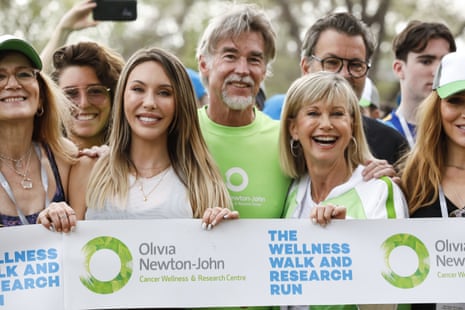 Chloe Lattanzi, John Easterling and Olivia Newton-John are smiling at the camera and holding up a banner with the logo of Olivia Newton-John Wellness Walk and Research Run and of the cancer centre. They are surrounded by other people at the event