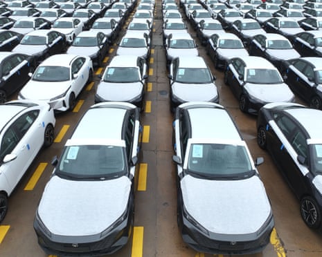Rows of white cars with yellow wheel blocks are parked in a large lot