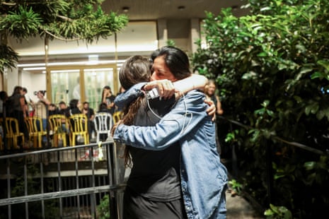 two people embrace outside the entrance to a building where people are gathered and chairs are lined up in front of a large window.