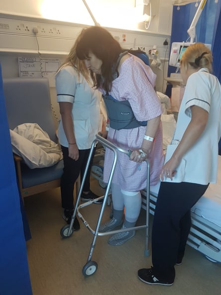 A woman using a zimmer frame in the hospital with physical therapists standing on either side of her.