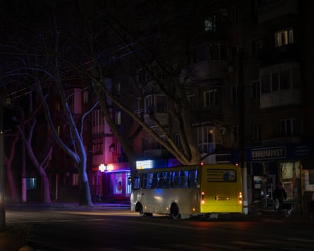 A bus makes its way along a dark street, the only light coming from a sign outside a pharmacy